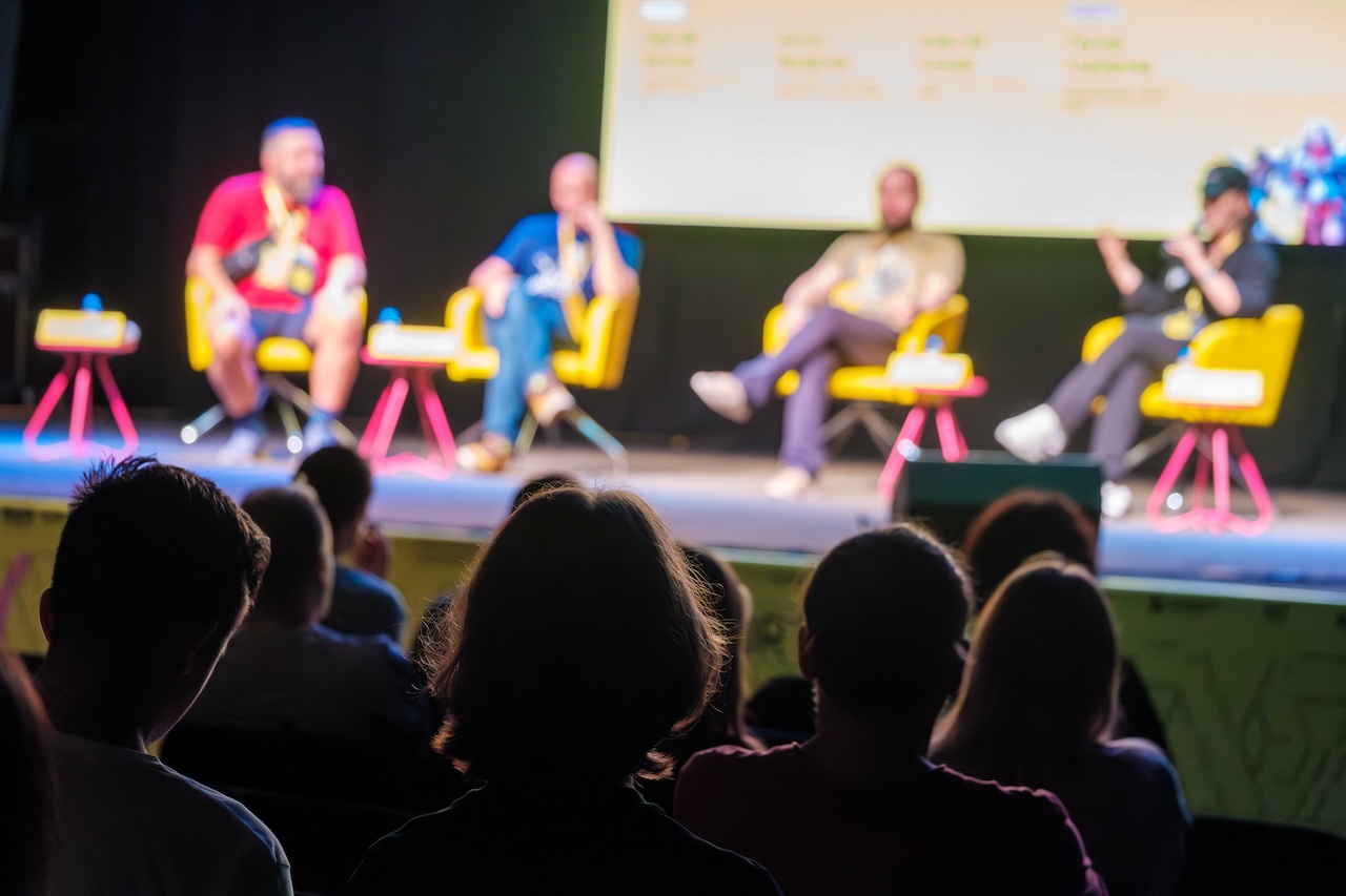 Colleagues sitting on stage during conference answering questions from audience in dark hall while presenting new project
